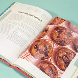 The Crumbs & Doilies Recipe Book open to chewy choc chip cookies with full color photos and baking tips.